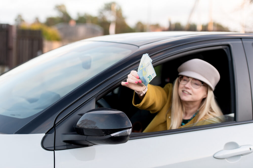 A woman wearing a beige hat and glasses leans out of a car window, holding several euro banknotes in her hand&mdash;perhaps celebrating savings made with advanced Transportation Management Software.