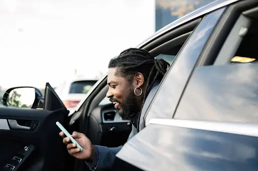 A person with dreadlocks and an earring sits in a car with the door open, smiling while looking at a smartphone&mdash;perhaps checking ride details on PerGO NEMT Software.