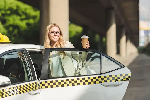 A smiling woman with glasses stands next to an open taxi door, holding a coffee cup and a white bag, showcasing how PerGO NEMT Software streamlines transportation management in a bustling city environment surrounded by greenery.