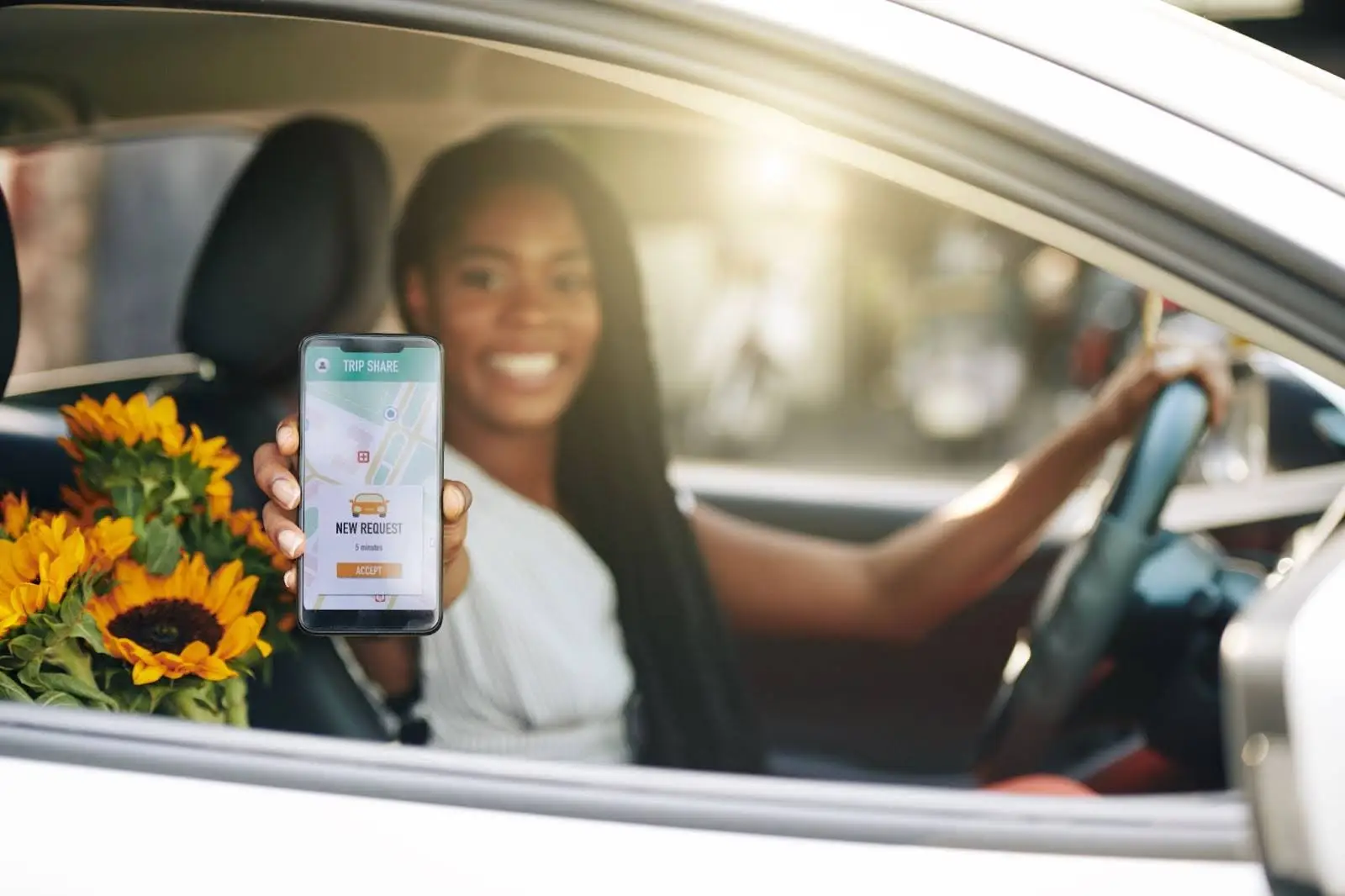 A smiling woman sits in a car&rsquo;s driver seat, holding a smartphone with a ride-sharing app on the screen. Sunflowers rest on the passenger seat beside her. Sunlight shines through the car window.