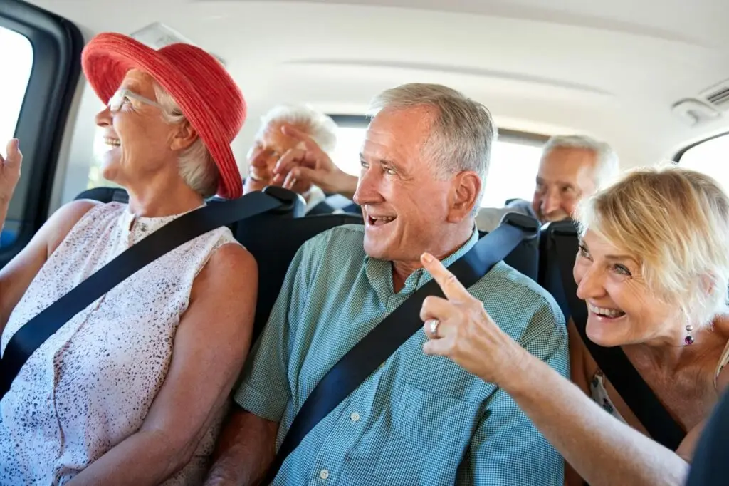 A group of smiling older adults, wearing seatbelts, sit in the backseat of a car. They appear to be laughing and talking, enjoying each others company. One woman in front is pointing outside the window.