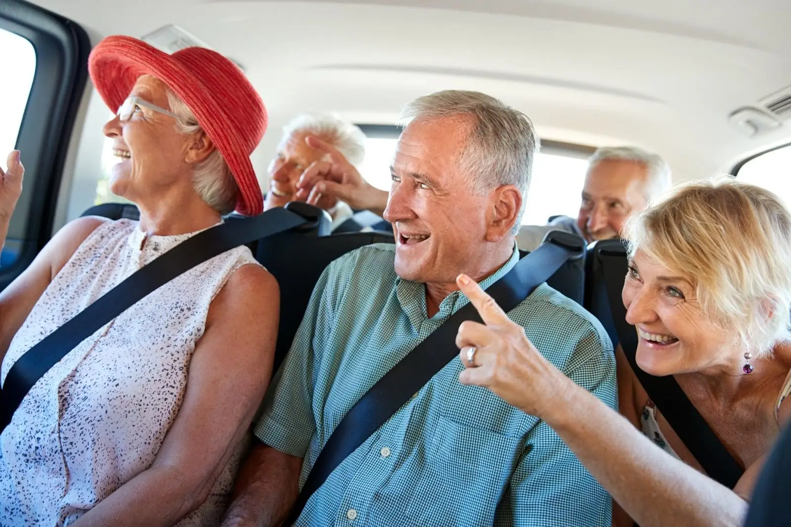 A group of smiling older adults, wearing seatbelts, sit in the backseat of a car. They appear to be laughing and talking, enjoying each others company. One woman in front is pointing outside the window.