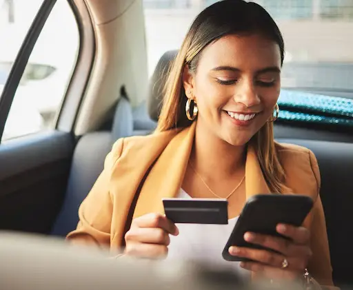A woman sitting in a car smiles while holding a credit card and using her smartphone, possibly making an online purchase or mobile payment.