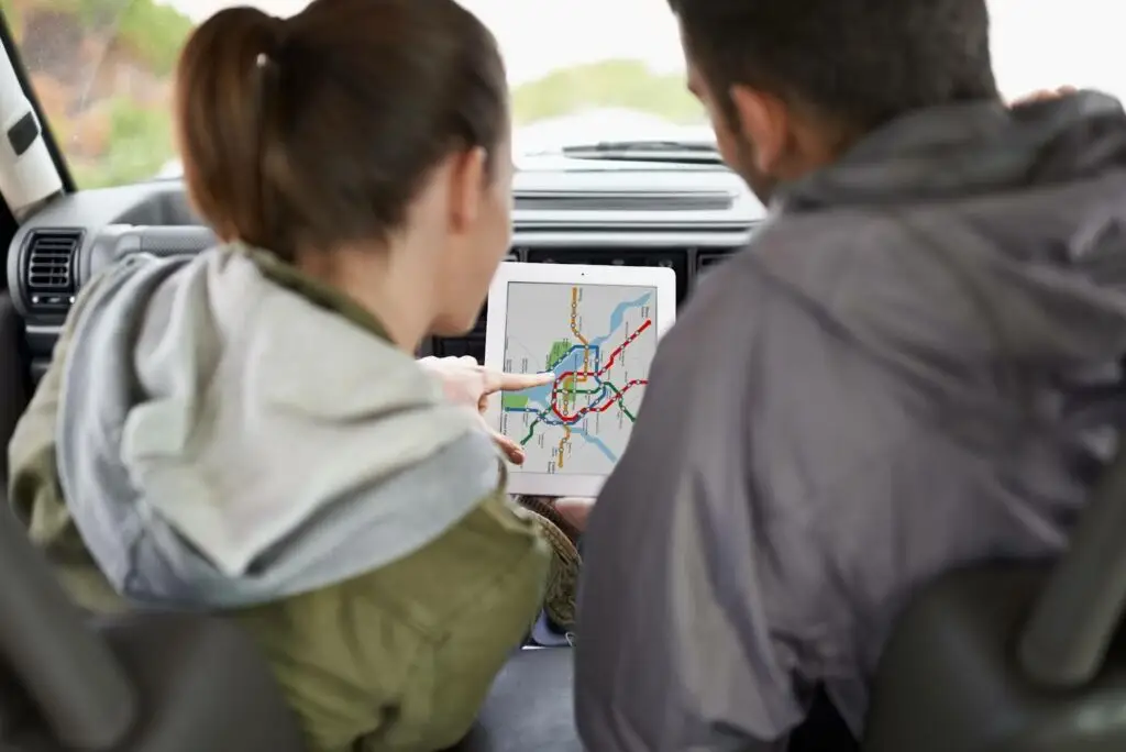 Two people sitting in a car look at a tablet displaying a city metro map, discussing directions or planning a route. Their faces are turned away from the camera, focusing on the screen.