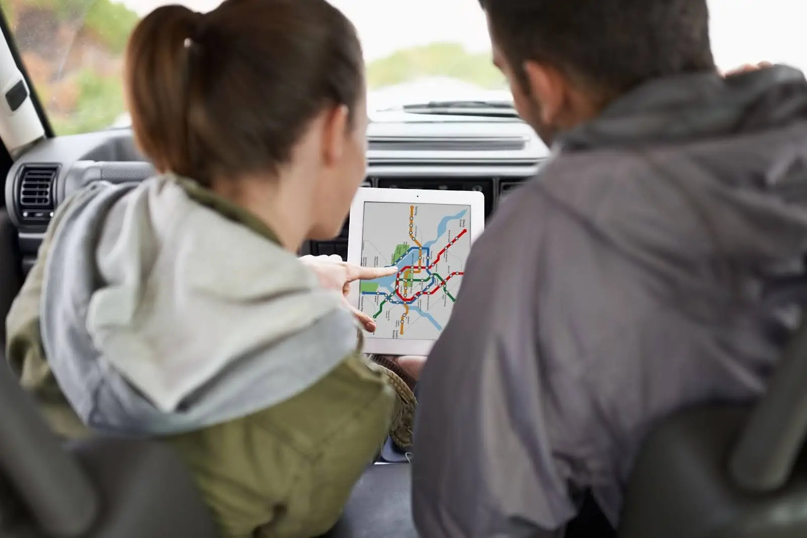 Two people sitting in a car look at a tablet displaying a city metro map, discussing directions or planning a route. Their faces are turned away from the camera, focusing on the screen.