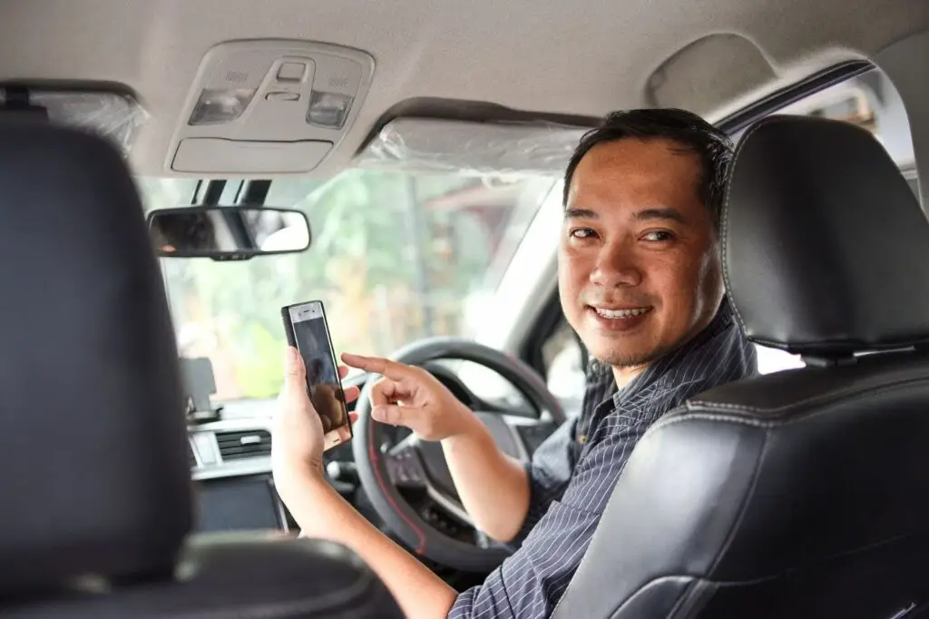 A smiling man sits in the driver’s seat of a car, holding a smartphone and pointing at its screen while looking back toward the camera.