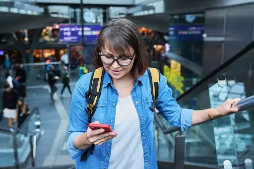A young woman with glasses and a backpack looks at her phone while riding an escalator in a busy, modern indoor public space, possibly a train station or airport.