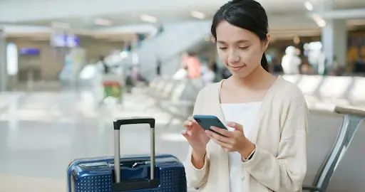A woman stands in an airport terminal beside a blue suitcase, looking at her smartphone. She is wearing a light cardigan and has her hair tied back. The background shows empty seats and blurred travelers.