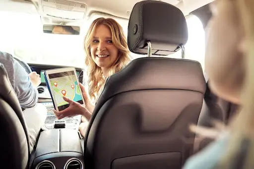 A woman sitting in the front passenger seat of a car smiles while holding a tablet displaying a map with location pins. The photo is taken from the back seat, and other passengers are partially visible.
