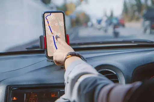 A person sitting in a car uses their finger to touch a smartphone mounted on the dashboard, displaying a GPS navigation map with a highlighted route. The car is driving on a sunny road.