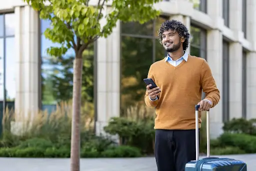 A smiling man with curly hair, wearing a brown sweater over a blue shirt, stands outdoors holding a smartphone in one hand and the handle of a blue suitcase in the other, near a modern building and greenery.