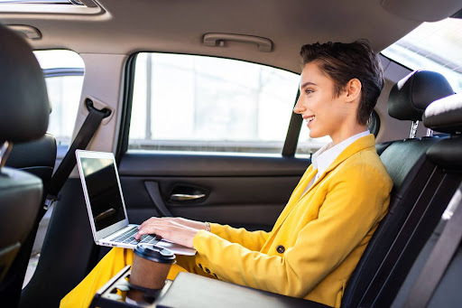 A person in a bright yellow suit sits in the backseat of a car, smiling while working on a laptop. A coffee cup is in the car&rsquo;s center armrest holder. Sunlight streams through the window.