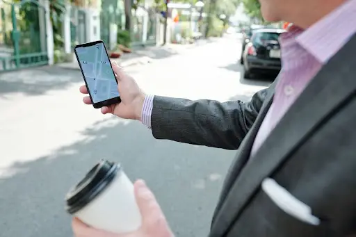 A person in a suit stands on a street holding a cup of coffee and looking at a smartphone displaying a map app with route directions.