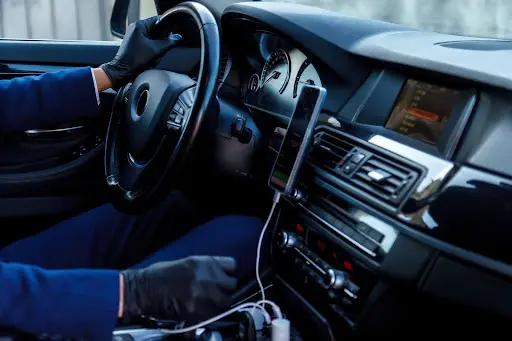 A person wearing black gloves is driving a car. The dashboard displays instruments, a touchscreen, and a smartphone connected with a charging cable&mdash;showcasing the modern interior often seen in vehicles using Transportation Management Software.