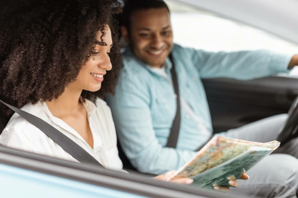 Smiling woman in the passenger seat reads a map while a man drives, both wearing seatbelts inside a car—an ideal scene for efficient routes managed with Transportation Dispatch Software.