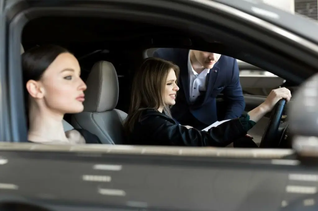 Two women sit in the front seats of a car, with one in the driver’s seat holding the steering wheel and smiling. A man standing outside leans in, engaging with them—showcasing seamless trips powered by PerGO NEMT Software.