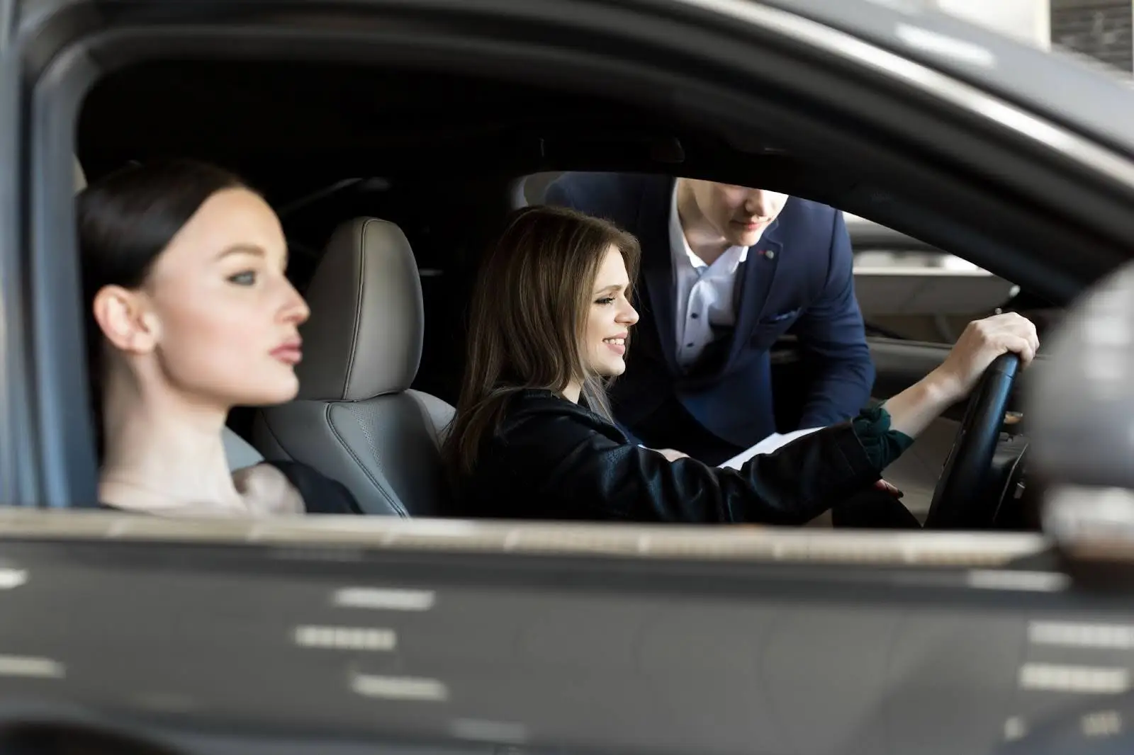 Two women sit in the front seats of a car, with one in the driver&rsquo;s seat holding the steering wheel and smiling. A man standing outside leans in, engaging with them&mdash;showcasing seamless trips powered by PerGO NEMT Software.