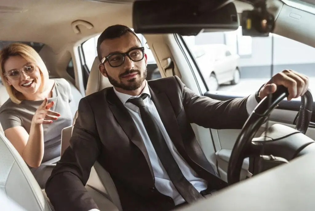 A man in a suit and glasses sits in the driver’s seat of a car, holding the steering wheel. A smiling woman in the back seat leans forward and points, appearing to talk to the driver—perhaps discussing their new Transportation Dispatch Software.