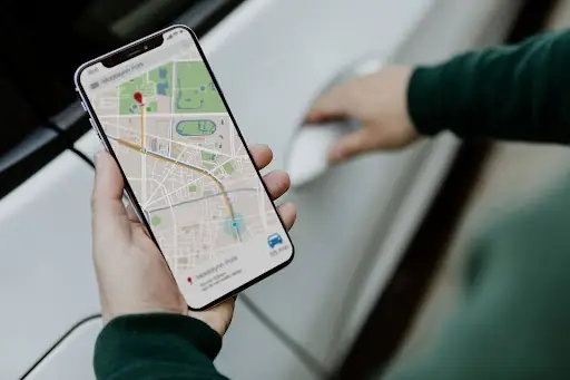 A person holding a smartphone displaying a navigation map while reaching to open a white car door, showcasing how Transportation Management Software streamlines trip coordination on the go.