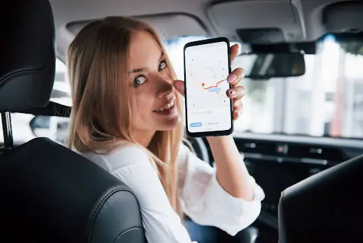 A woman sitting in a car's driver seat smiles and holds up a smartphone displaying NEMT Software with a map on the screen, ready for her next ride.