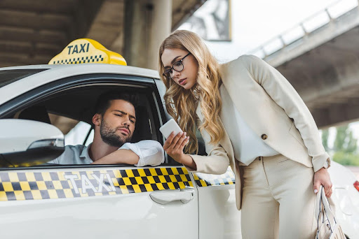 A woman in a beige suit shows her phone, featuring PerGO NEMT Software, to a male taxi driver leaning out of the window under an overpass. The driver looks at the screen with a serious expression.