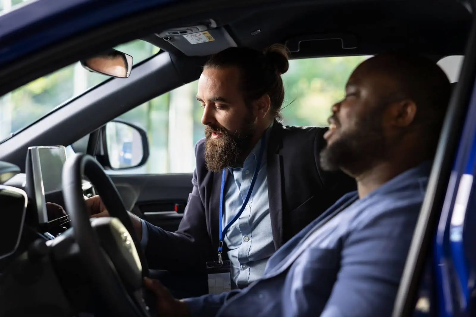 Two men sit in the front seats of a car. One points at the dashboard display, explaining features of PerGO NEMT Software to the driver. Both appear engaged in conversation inside a modern vehicle equipped with transportation management software.