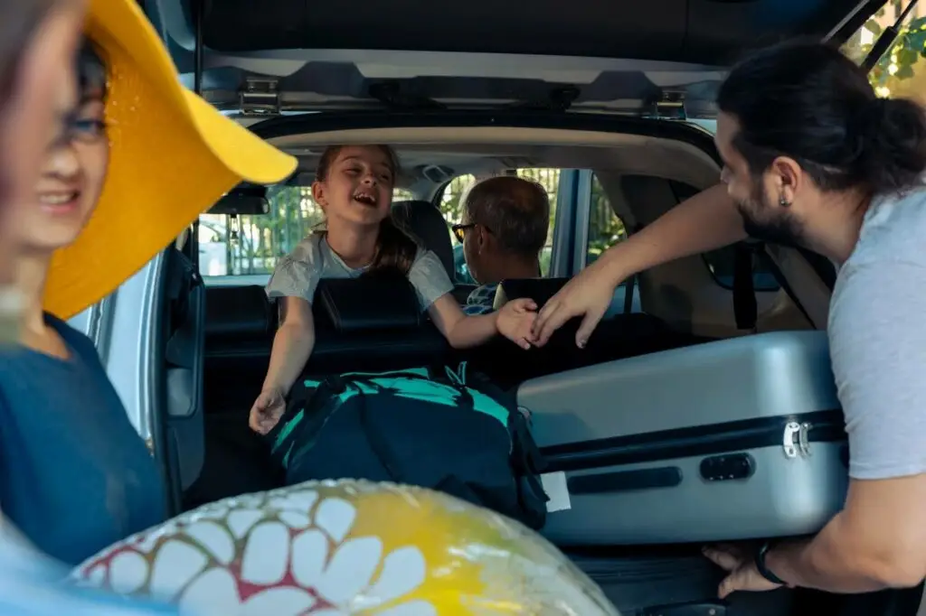 A family packs their car for a trip. A smiling girl sits in the back seat, reaching out, while adults load luggage and a beach ball into the trunk—just like a seamless journey managed with PerGO NEMT Software. Sunlight filters through the trees.