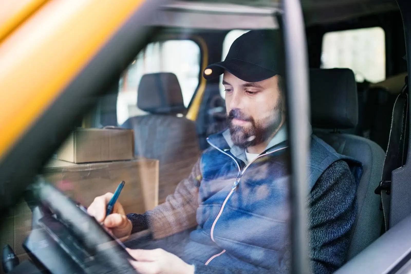 A bearded man wearing a cap sits in the driver&rsquo;s seat of a vehicle, writing on a notepad with PerGo NEMT Software as cardboard boxes are stacked in the back. The photo is taken through the window.