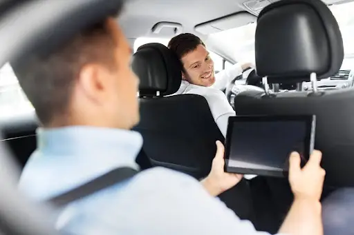 A man in the back seat of a car holds a tablet while another man in the front seat turns around, smiling and talking to him. Both appear to be having a friendly conversation during a car ride.
