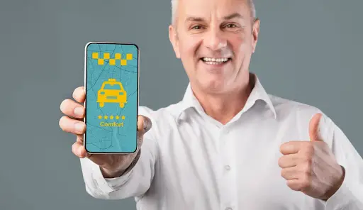 Smiling man in a white shirt holds up a smartphone displaying a taxi app with a yellow taxi icon and gives a thumbs up, suggesting satisfaction with the service.