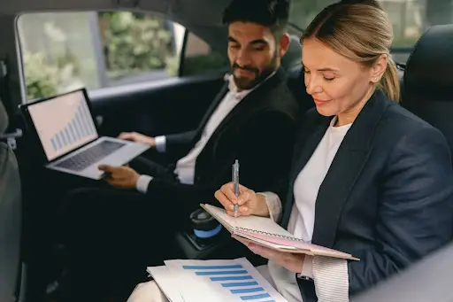 Two business professionals sit in a car; the man uses a laptop displaying charts, while the woman writes in a notebook, with printed graphs on her lap. Both are dressed in business attire.