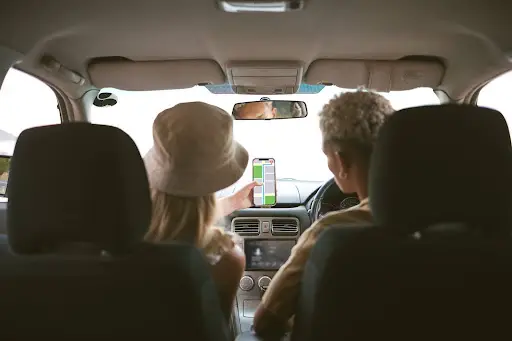 Two people sit in the front seats of a car, with the passenger holding up a smartphone displaying a navigation app to help guide the driver. The photo is taken from the back seat.
