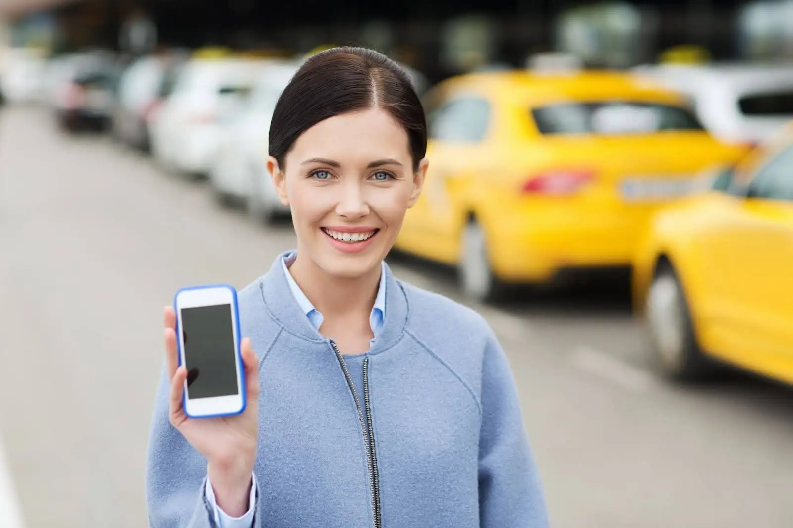 A smiling woman stands outdoors near a row of yellow taxis, holding up a smartphone with a blank screen, wearing a light blue coat.