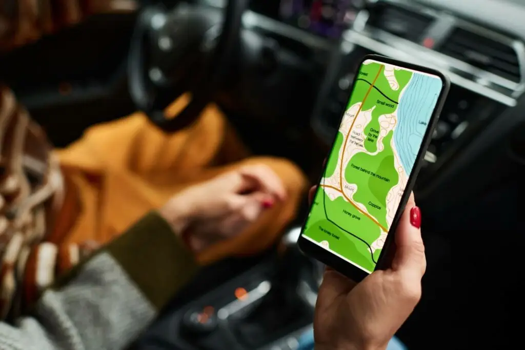 A person sits in a car holding a smartphone displaying a colorful map with green land areas, roads, and blue water, using navigation while the car is parked.