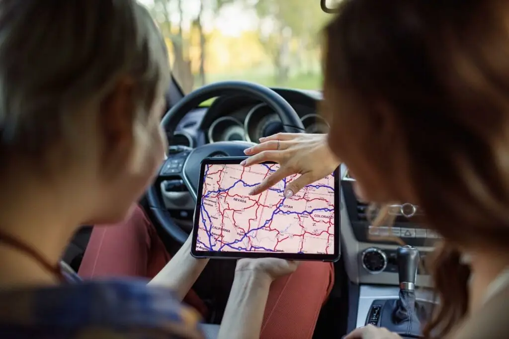 Two people sitting in a car look at a tablet displaying a detailed road map. One person points at a location on the screen, while the other holds the tablet near the steering wheel.