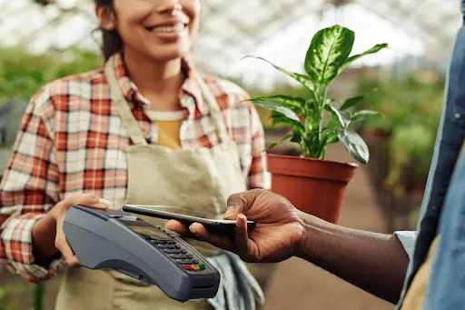 A person holds a smartphone over a card reader to make a contactless payment, while a smiling worker in an apron holds a potted plant in the background at a greenhouse or plant shop.