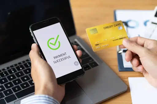 A person holding a smartphone displaying a &ldquo;Payment Successful&rdquo; message with a green checkmark in one hand and a yellow credit card in the other, next to a laptop and office supplies on a desk.