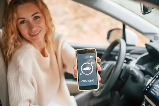 A woman sitting in a car smiles and holds up a smartphone displaying a car charging screen, indicating the vehicle is currently charging.
