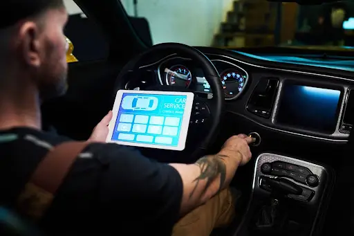 A man sits in a car holding a tablet with a Car Service screen displayed, while turning the ignition key. The car dashboard and controls are visible, indicating diagnostics or maintenance.