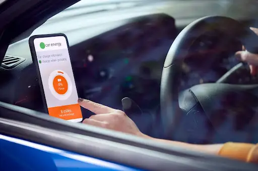 A person in a car uses a smartphone mounted on the dashboard, displaying an app that shows electric car charging status and range in miles. The drivers hand is about to tap the screen.