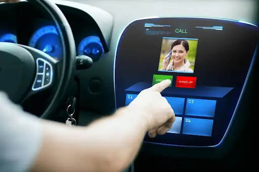 A person&rsquo;s hand is about to press the ANSWER button on a car&rsquo;s touchscreen display, which shows an incoming video call with a smiling woman on the screen. Car dashboard and steering wheel are visible.