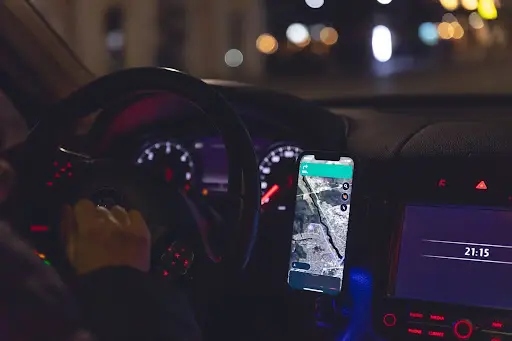 A person drives a car at night with a smartphone showing a navigation app mounted on the dashboard and the car clock displaying 21:15. The dashboard lights and city streetlights are visible.