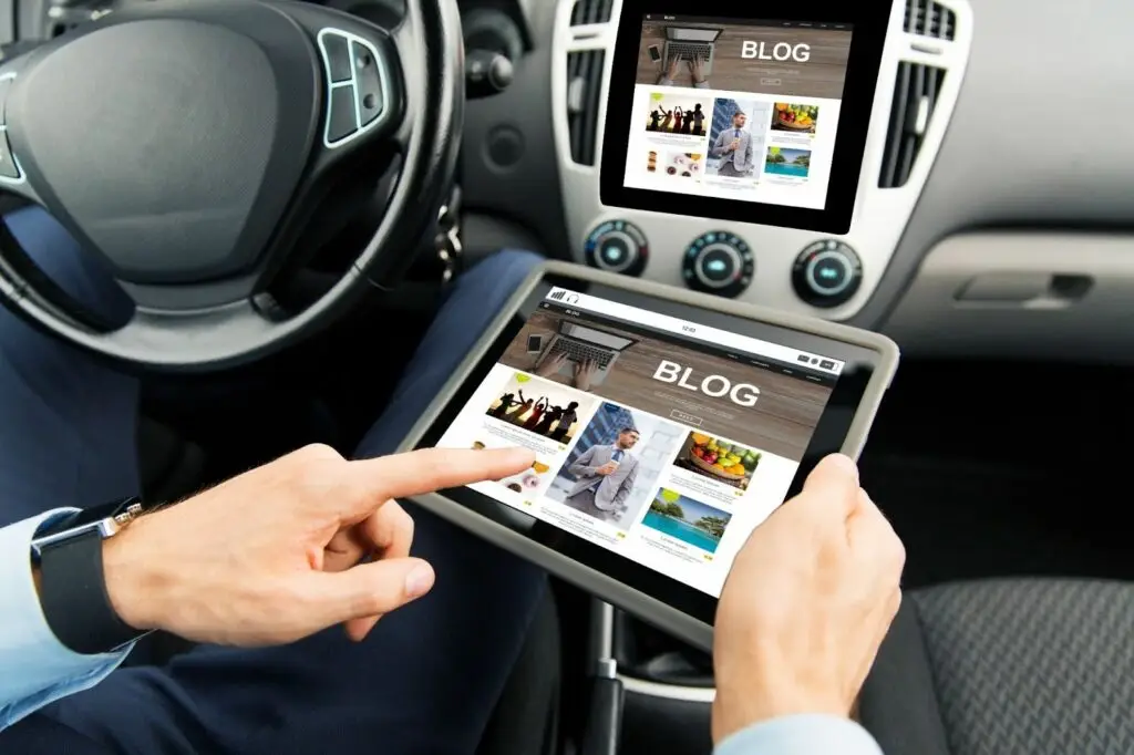 A person in a car uses a tablet displaying a blog webpage, with the same blog shown on the car’s dashboard screen. The car interior and steering wheel are visible.