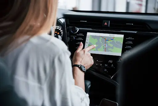A person in the driver&rsquo;s seat uses a touchscreen car navigation system, pointing at a map displayed on the screen inside a modern vehicle.