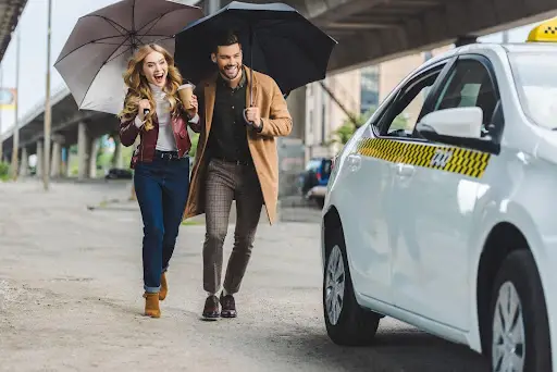 A cheerful woman and man holding umbrellas are running toward a white taxi parked on a city street, appearing excited and ready to get in.