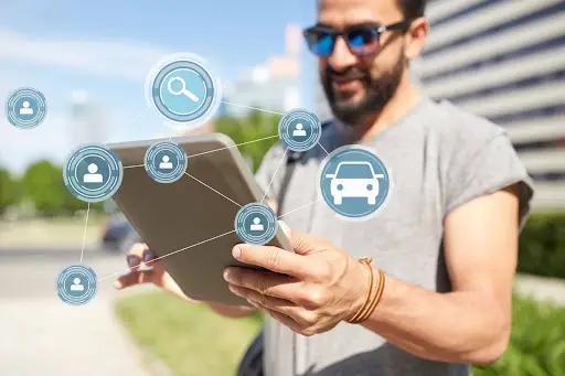 A man wearing sunglasses uses a tablet outdoors. Digital icons for users, a car, and a magnifying glass are overlaid, suggesting concepts like networking, search, and advanced Transportation management Software such as PerGo.