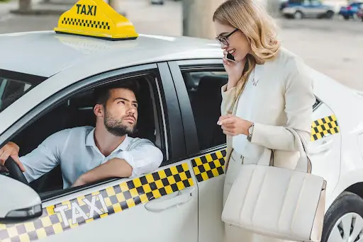 A man driving a taxi looks out the window at a woman standing beside the car, who is talking on her phone and holding a large white handbag, possibly using PerGo transportation dispatch software to arrange her ride.