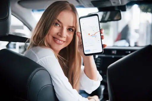 Smiling woman sitting in a car shows a smartphone with PerGo NEMT Software open, displaying a navigation route for seamless transportation dispatch.