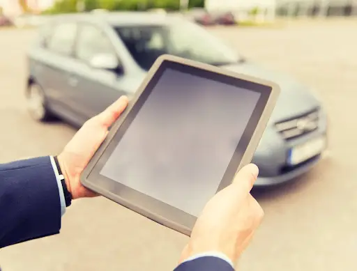 A person in a suit holds a tablet outdoors with both hands, standing near a gray car parked on a paved surface. The blank tablet screen hints at using transportation dispatch software like PerGo, with the background slightly blurred.