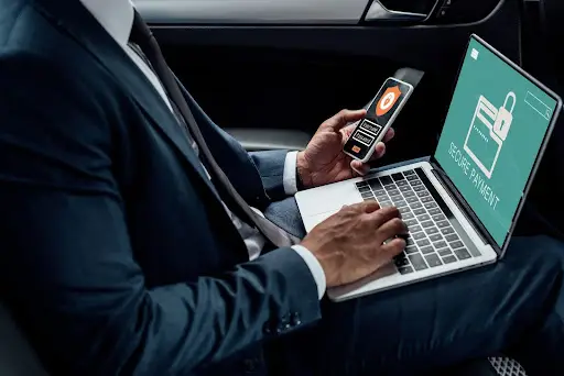 A person in a suit sits in a car using a laptop with a &ldquo;Secure Payment&rdquo; screen and holds a smartphone displaying a Bitcoin symbol, suggesting online cryptocurrency transactions, possibly managed through PerGo NEMT Software.
